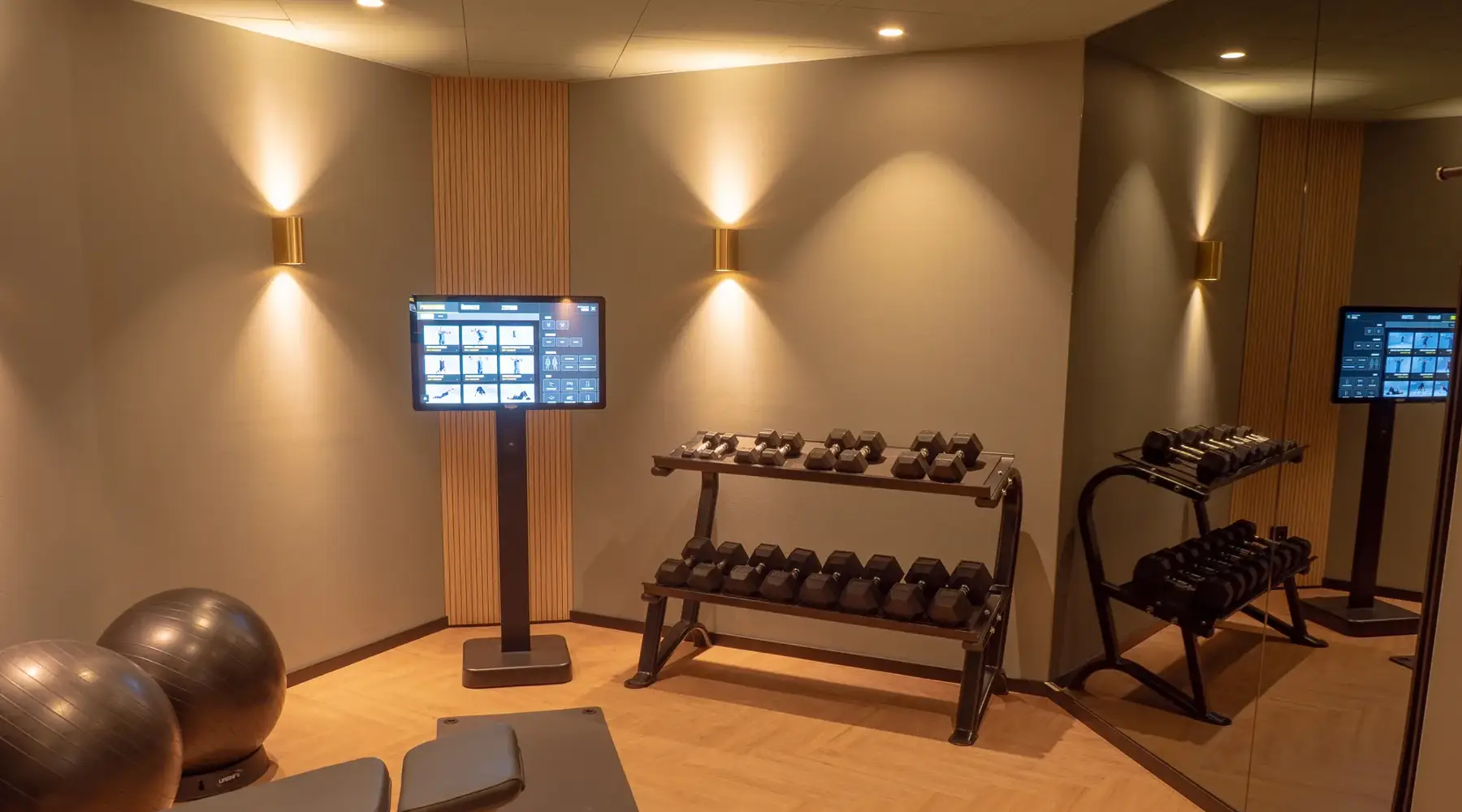 Fitnessbereich im Van der Valk Hotel Hildesheim mit Hanteln auf einem mehrstufigen Rack, Gymnastikbällen und einem digitalen Trainingsdisplay. Der Raum ist modern gestaltet, mit Holzfußboden und indirekter Beleuchtung, und bietet eine funktionale Umgebung für Kraft- und Fitnesstraining.