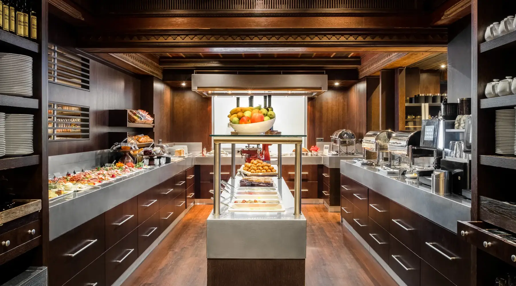 Modern gestalteter Buffetbereich im Van der Valk Hotel Hildesheim mit dunklen Holzfronten und hellen Arbeitsflächen. Links und rechts verlaufen lange Buffetstrecken mit einer Vielzahl an Speisen, darunter kalte Platten, Obst und weitere angerichtete Spezialitäten. In der Mitte steht eine beleuchtete Insel mit Desserts und Gebäck, darüber eine Schale mit frischem Obst. Auf der rechten Seite befinden sich Kaffeemaschinen und Warmhaltegeräte, während die warme Beleuchtung und die hochwertige Ausstattung eine einladende, gepflegte Atmosphäre schaffen.