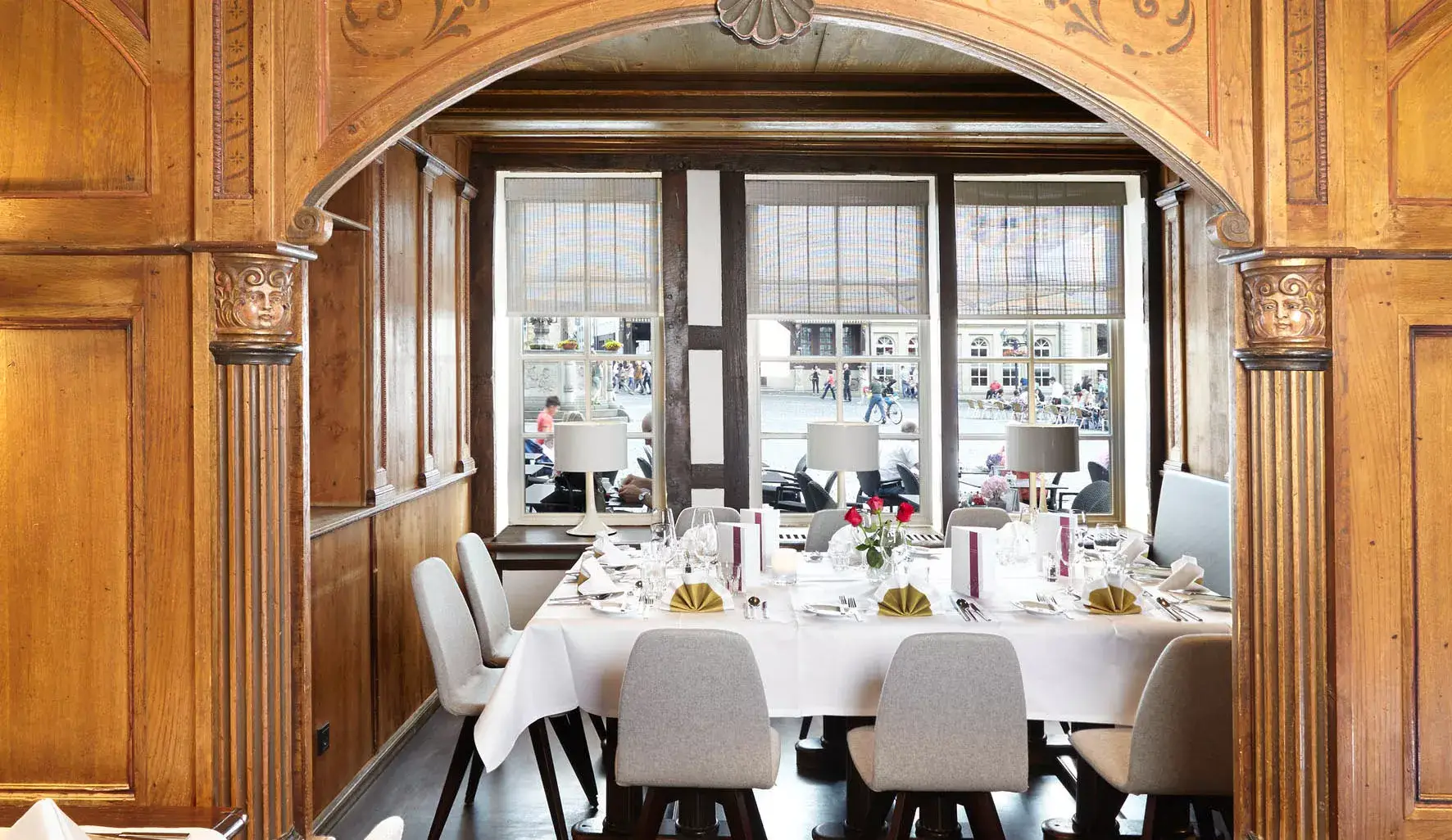 Blick in den hellen Restaurantbereich des Van der Valk Hotel Hildesheim mit einer festlich eingedeckten Tafel vor großen Fenstern. Der Raum ist von historischen Holzelementen, verzierten Säulen und einer warmen Holzvertäfelung geprägt. Auf dem weiß gedeckten Tisch stehen Gläser, Besteck, Menükarten, gefaltete goldene Servietten und eine Vase mit roten Rosen. Durch die Fenster fällt Tageslicht ein, im Hintergrund ist der Marktplatz zu erkennen.