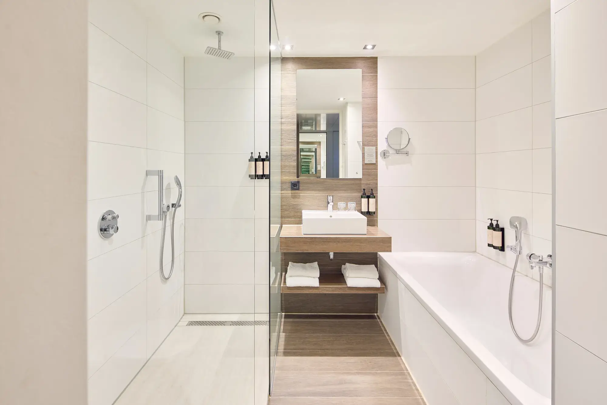 Modern hotel bathroom with a spacious walk-in shower, bathtub, washbasin, large mirror, and light wall tiles with wood accents.