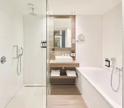 Modern hotel bathroom with a spacious walk-in shower, bathtub, washbasin, large mirror, and light wall tiles with wood accents.