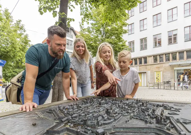 Four-member family stands outdoors around a detailed city model made of metal and looks at it curiously; two children point to specific places while the parents bend over it with interest, surrounded by trees and buildings in a quiet urban environment.
