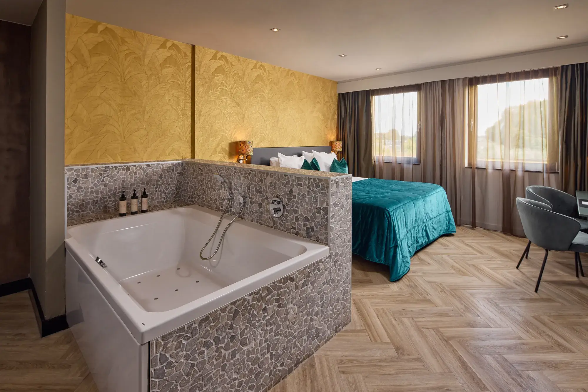 Luxe hotelsuite met bubbelbad, tweepersoonsbed met turquoise bedsprei, goudkleurig behang met botanisch patroon, visgraatvloer en zithoek bij de ramen.