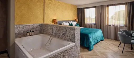 Luxe hotelsuite met bubbelbad, tweepersoonsbed met turquoise bedsprei, goudkleurig behang met botanisch patroon, visgraatvloer en zithoek bij de ramen.