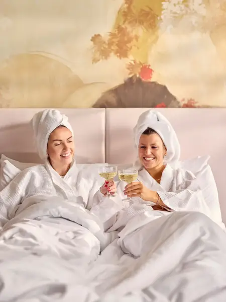 Two women in bathrobes enjoying a drink on a bed in a hotel room at Van der Valk Hotel Ghent, with towels and a warm, relaxing atmosphere.