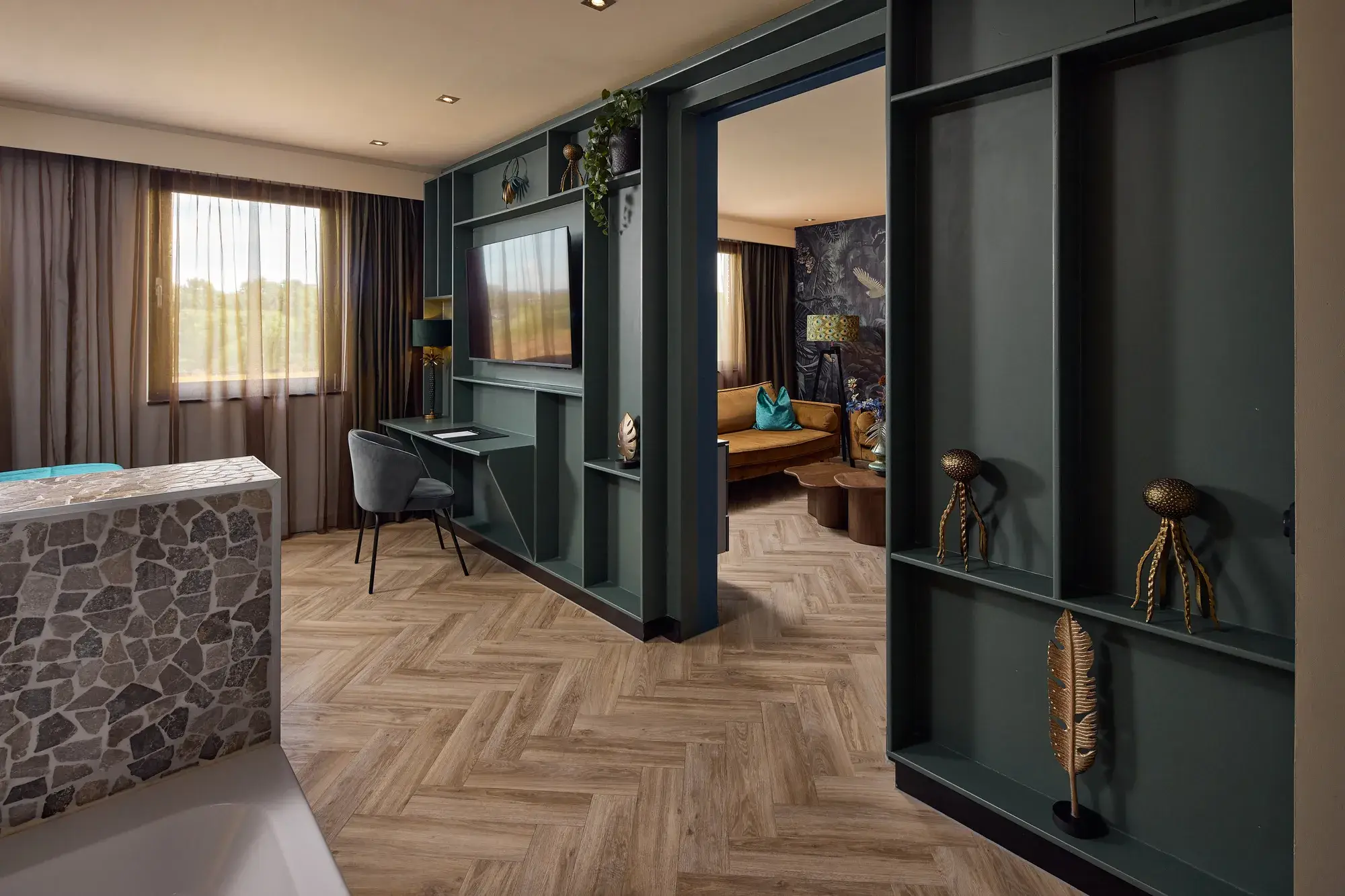 Luxe hotelsuite met donkergroene wandkast, televisie, bureau, visgraatvloer en doorgang naar een aparte zithoek met bank.