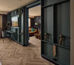 Luxe hotelsuite met donkergroene wandkast, televisie, bureau, visgraatvloer en doorgang naar een aparte zithoek met bank.