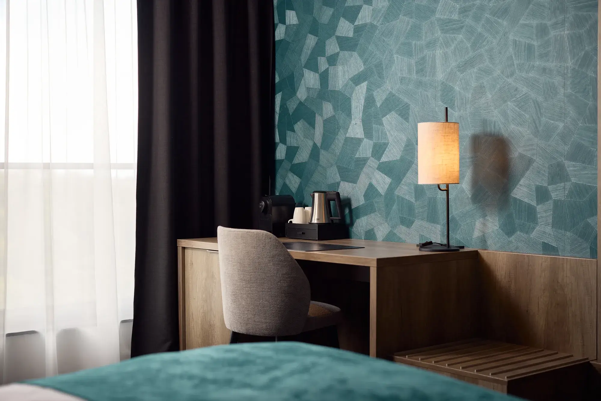 Hotel room with wooden desk, chair, coffee and tea making facilities, atmospheric table lamp, and turquoise wall covering with a geometric pattern.