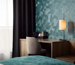 Hotel room with wooden desk, chair, coffee and tea making facilities, atmospheric table lamp, and turquoise wall covering with a geometric pattern.