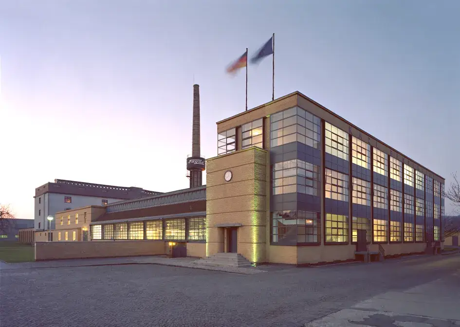 Exterior view of a modern industrial building with large glass facades and clear, functional architecture. The building is multi-story, with rectangular shapes and illuminated interiors visible through the windows. In the background, a tall chimney rises, while the building stands in an open setting with a driveway and free space in front.