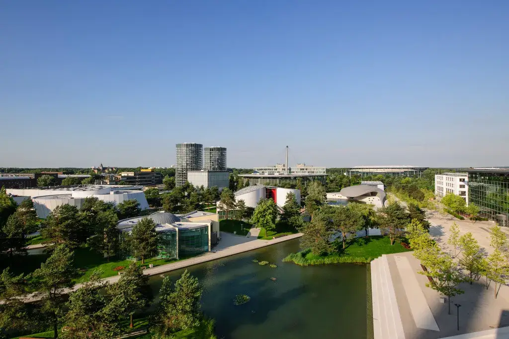 Luftaufnahme eines modernen Stadtgebiets mit mehreren architektonisch auffälligen Gebäuden, darunter runde und futuristisch gestaltete Bauwerke, umgeben von Grünflächen, Bäumen und Wasserläufen. Im Hintergrund sind weitere Gebäude und Hochhäuser zu sehen, während im Vordergrund ein angelegter Park mit Wegen und einem kleinen Gewässer verläuft.