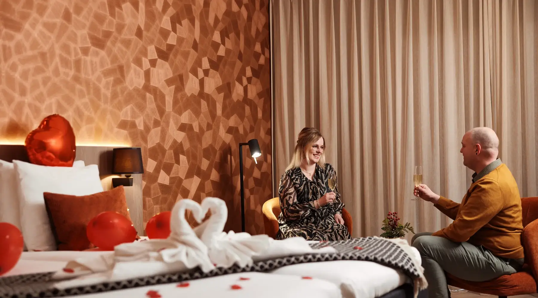 man and woman enjoying champagne with heart-shaped swan on bed
