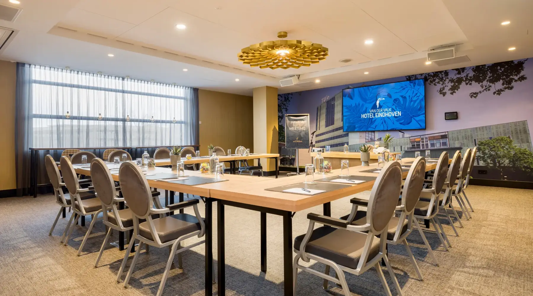 Salle à l'Hôtel Eindhoven. Les chaises et les tables sont disposées en forme de U. Au début de la salle, contre le mur, il y a un écran.