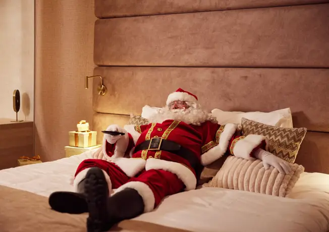 kerstman ligt in hotelbed met afstandsbediening