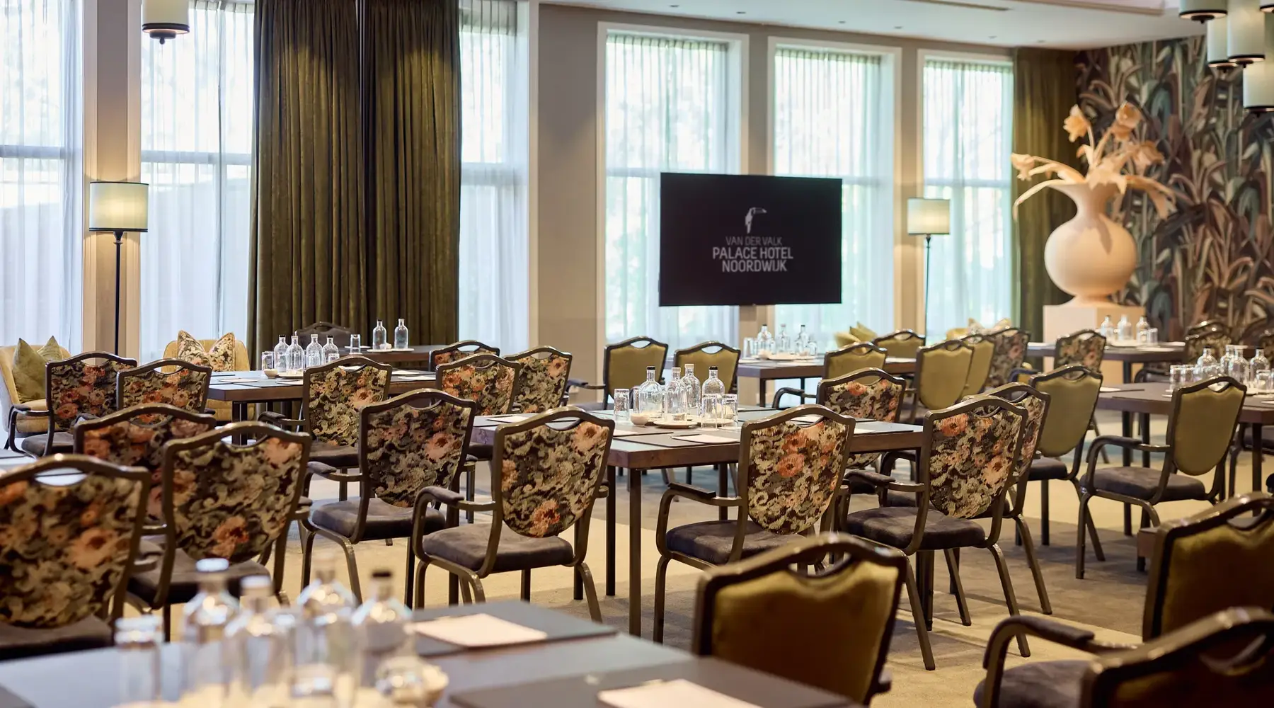 Salle de réunion Palace Hotel Noordwijk avec des chaises fleuries, des tables brunes et une télévision à l'avant de la salle.