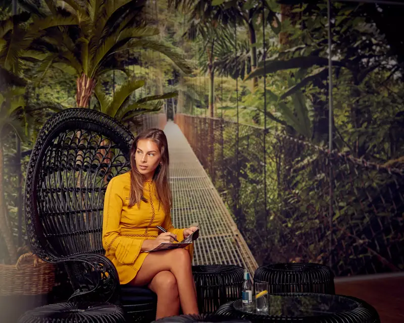 Een vrouw in een gele jurk zit in een rieten stoel met een notitieboek, in een tropisch ingerichte ruimte met jungle-achtergrond.