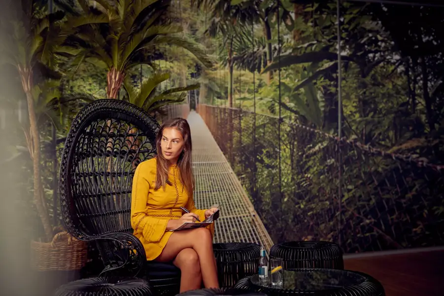 Een vrouw in een gele jurk zit in een rieten stoel met een notitieboek, in een tropisch ingerichte ruimte met jungle-achtergrond.