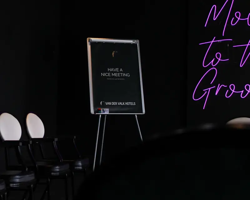 Stoelen in een rij met een flip-over waarop “Have a nice meeting” staat, in een donkere disco-vergaderruimte met neonletters.