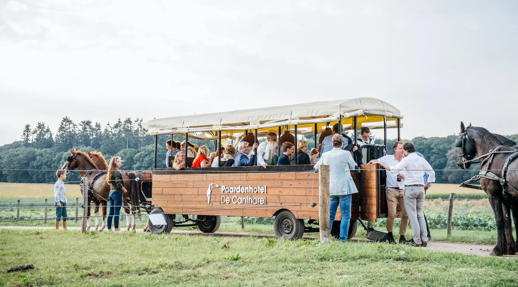 A covered wagon outside in the green in front of the hotel with 4 horses and many people who are just dressed and holding an event here. 
