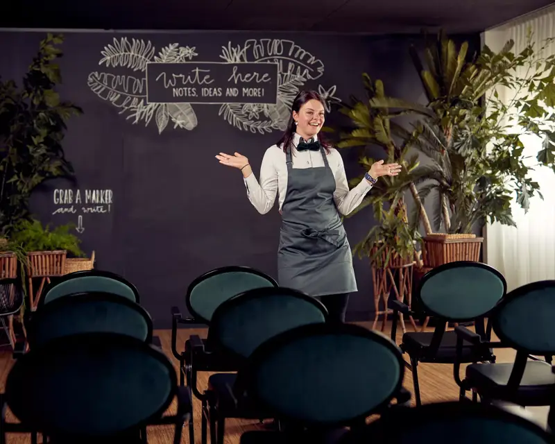 Een vrouw in een grijs schort staat lachend voor een zaal met stoelen, met achter haar een grote krijtwand en veel planten.
