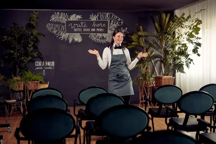 Een vrouw in een grijs schort staat lachend voor een zaal met stoelen, met achter haar een grote krijtwand en veel planten.