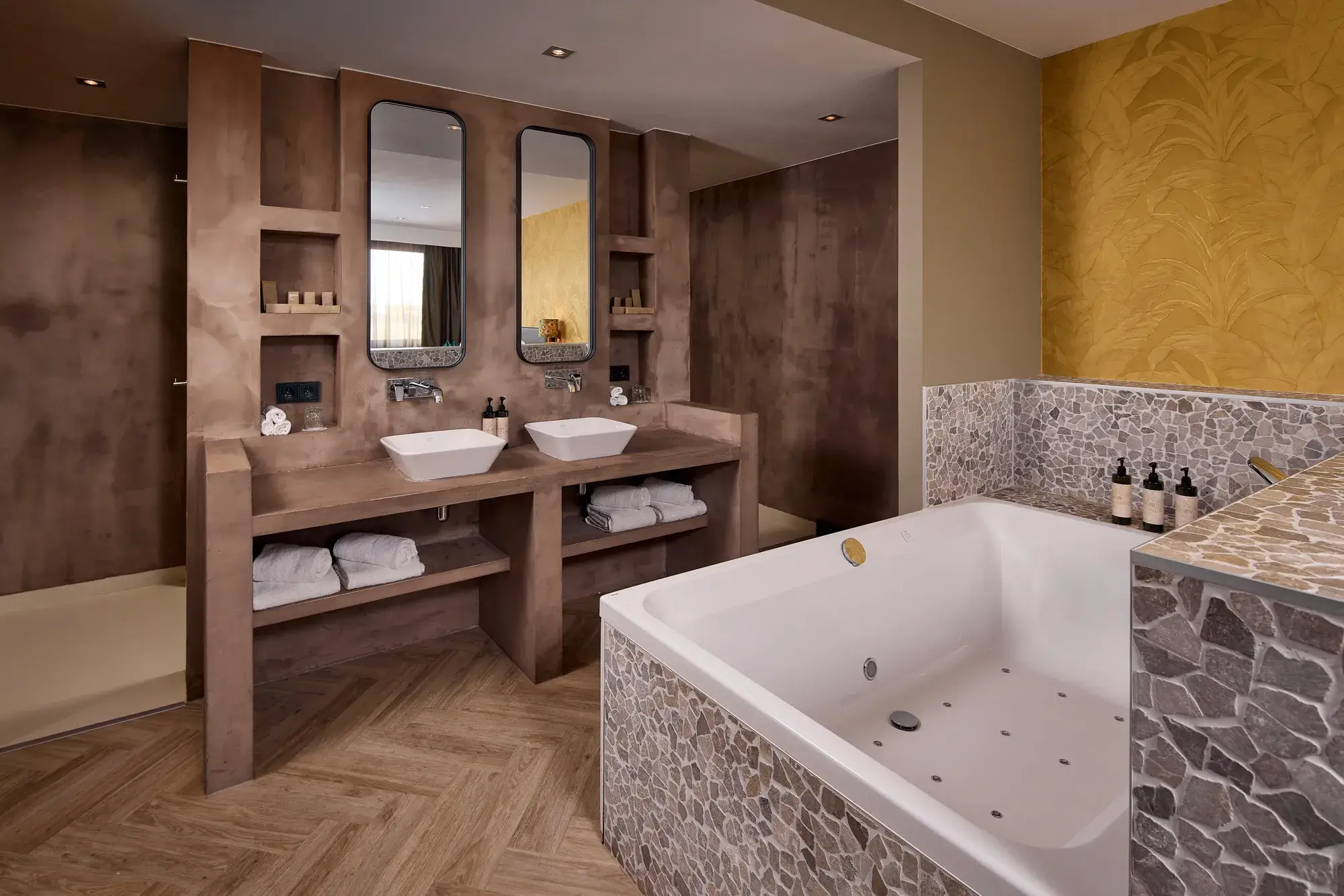 Luxe badkamer in hotelsuite met ruim bubbelbad, dubbele wastafel, twee spiegels, natuurstenen wandafwerking, inloopdouche en houten visgraatvloer.