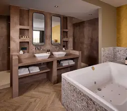Luxe badkamer in hotelsuite met ruim bubbelbad, dubbele wastafel, twee spiegels, natuurstenen wandafwerking, inloopdouche en houten visgraatvloer.