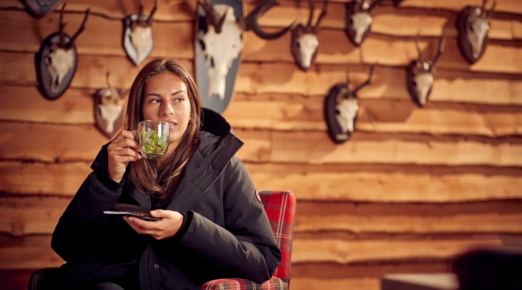 Eine Frau im warmen Mantel sitzt in einem Sessel mit einer Tasse Tee, vor einer Holzwand mit Jagdtrophäen.