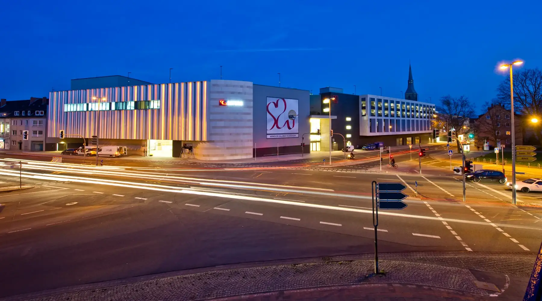 Exterior view of the Arneken Galerie in Hildesheim at dusk. The modern shopping center with its distinctive facade is brightly lit and forms a central point in the cityscape. In the foreground, roads with flowing traffic provide light trails that convey dynamism and urbanity. The combination of contemporary architecture and vibrant surroundings underscores the appeal of the Arneken Galerie as a popular destination for shopping and leisure.