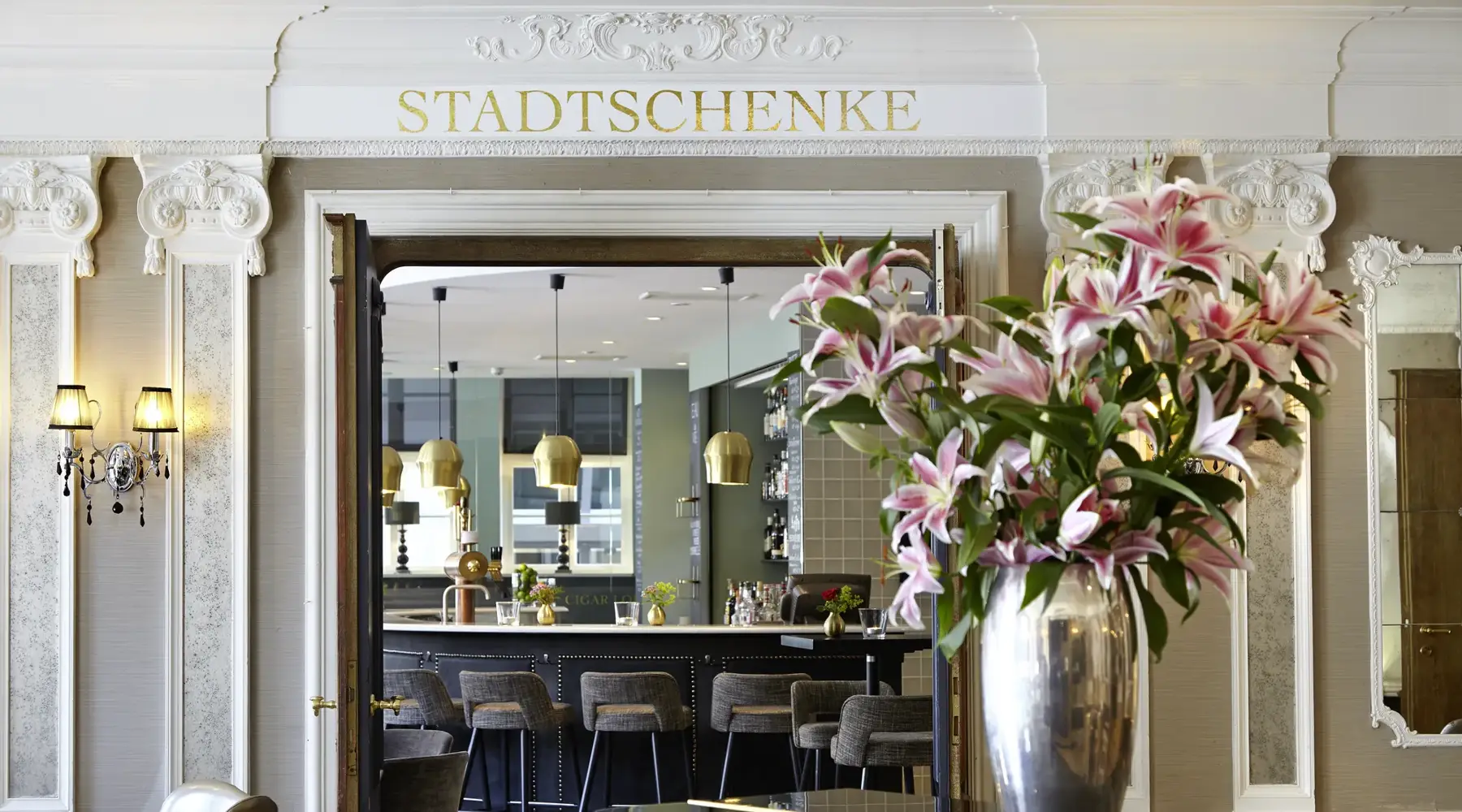 Blick durch den Rokoko-Eingang in die Stadtschenke des Van der Valk Hotels Hildesheim mit Bartresen und großem Blumenarrangement.