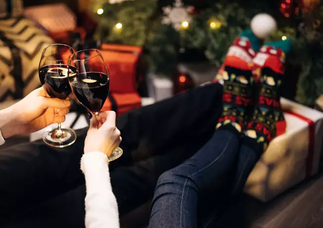 2 personen die gezellig samen liggen bij de kerstboom met cadeautjes, ze proosten met een glas rode wijn.