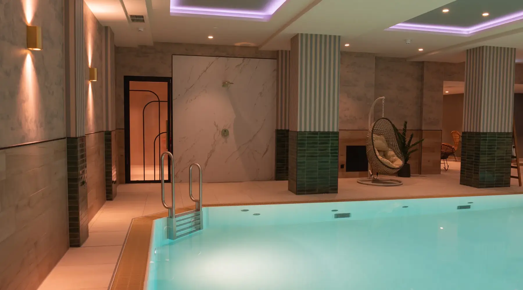 Indoor pool in the wellness area of the Van der Valk Hotel Hildesheim with illuminated water and modern design. At the edge of the pool is a metal entry staircase, in the background are tiled walls, a door to the sauna area, and a woven hanging chair can be seen. The warm lighting creates a calm, relaxing atmosphere.