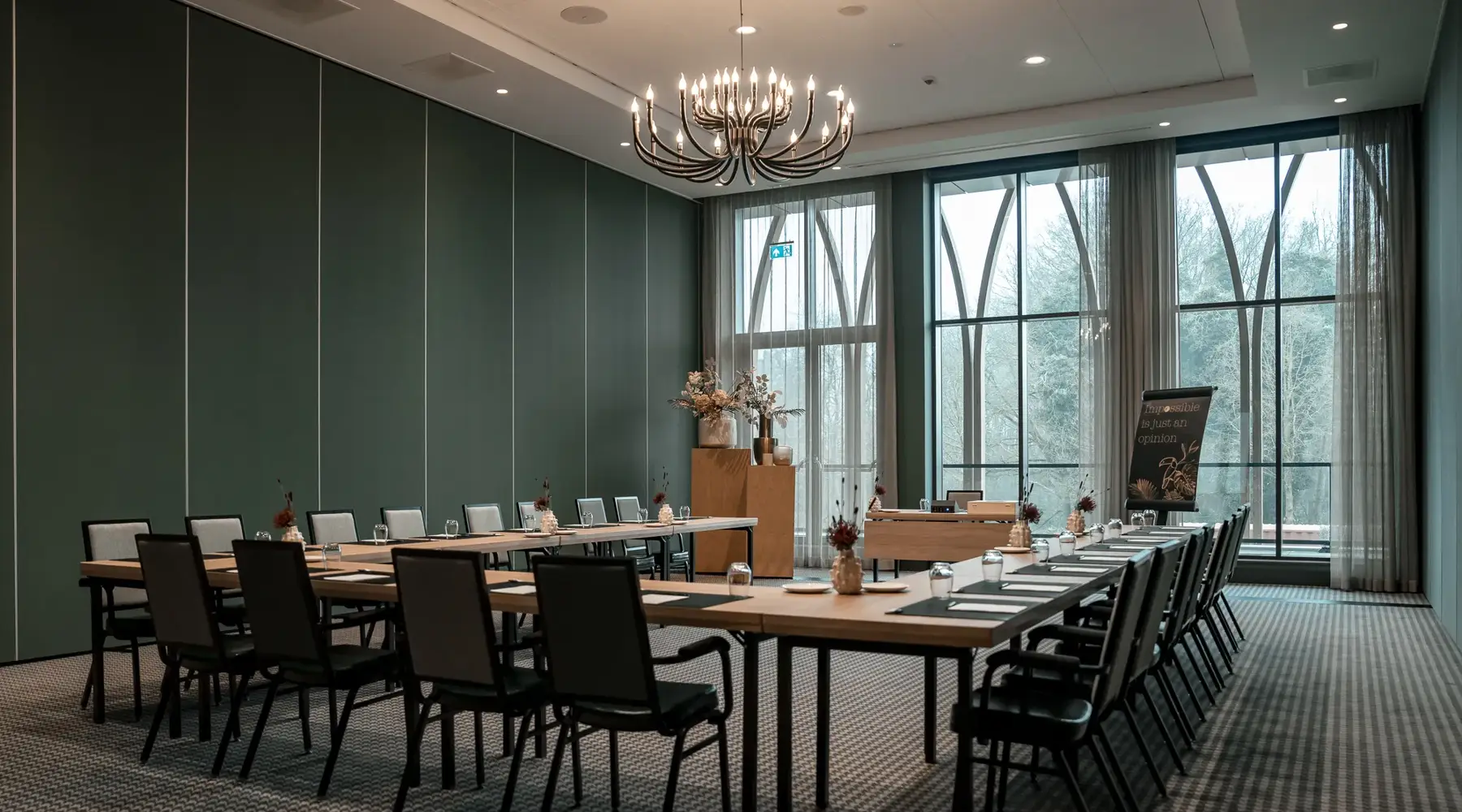 Zaal in Hotel Venlo. Tafels en stoelen staan in U-vorm. Zwart grijze stoelen staan achter de licht houten tafels.