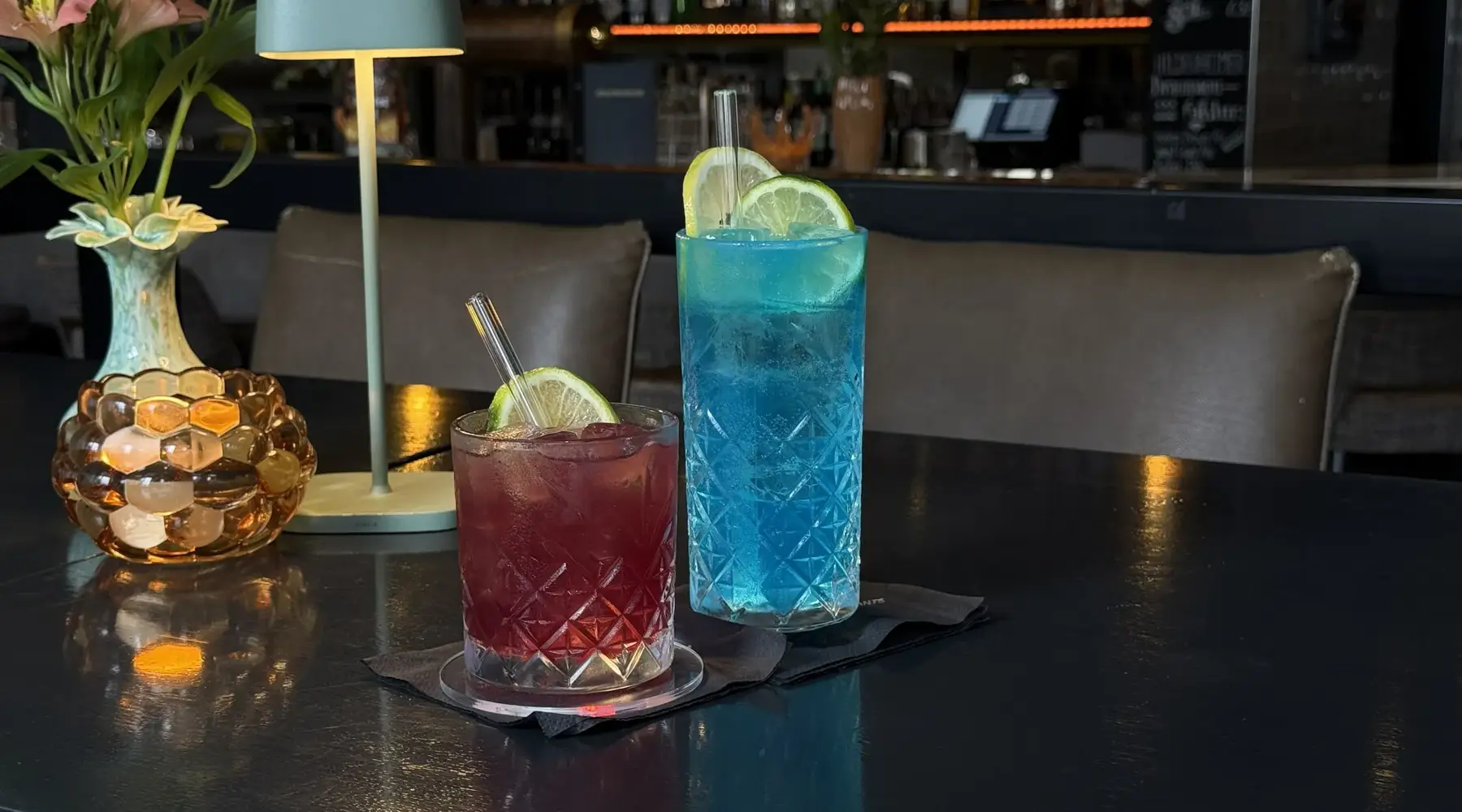 Op een donkere bartafel staan twee kleurrijke cocktails in stijlvolle geslepen glazen. De linker cocktail heeft een dieprode kleur, terwijl de rechter opvalt door zijn heldere blauwe tint. Beide drankjes worden geserveerd met ijs en verse schijfjes limoen. De sfeervolle verlichting en de moderne baromgeving zorgen voor een elegante en uitnodigende uitstraling.