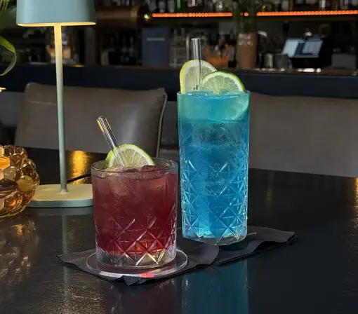 Zwei farbenfrohe Cocktails stehen in eleganten Kristallgläsern auf einem dunklen Bartisch. Der linke Cocktail hat eine tiefrote Farbe, während der rechte mit seinem leuchtenden Blau besticht. Beide Drinks werden mit Eis und frischen Limettenscheiben serviert. Die stimmungsvolle Beleuchtung und das moderne Bar-Ambiente schaffen eine elegante und einladende Atmosphäre.