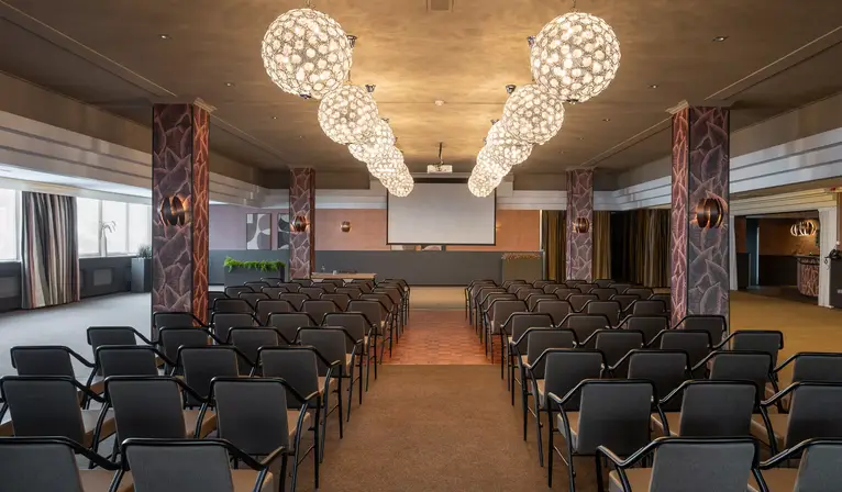 Overzicht van de ruime Valkenzaal ingericht in theateropstelling, voorzien van een groot presentatiescherm en opvallende bolvormige hanglampen.