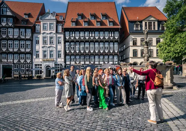 Geführte Stadtbesichtigung auf dem historischen Marktplatz in Hildesheim. Eine Gruppe von Teilnehmern folgt aufmerksam den Erläuterungen des Guides, während im Hintergrund die charakteristischen Fachwerkhäuser sowie das Van der Valk Hotel Hildesheim zu erkennen sind. Die Szene vermittelt die besondere Atmosphäre der Altstadt und verbindet kulturelle Entdeckung mit der zentralen Lage des Hotels.