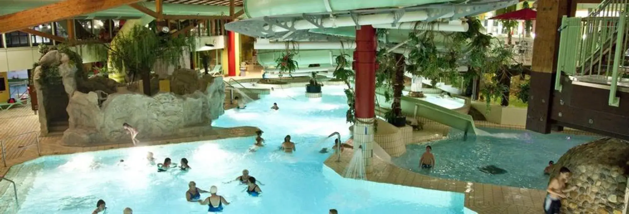 Binnengebied van een waterparadijs met meerdere zwembaden, warm water en tropisch ontworpen omgeving. Bezoekers ontspannen in het water, terwijl zuilen, planten en verschillende zwemgebieden een gevarieerde en ontspannende wellness-sfeer creëren.