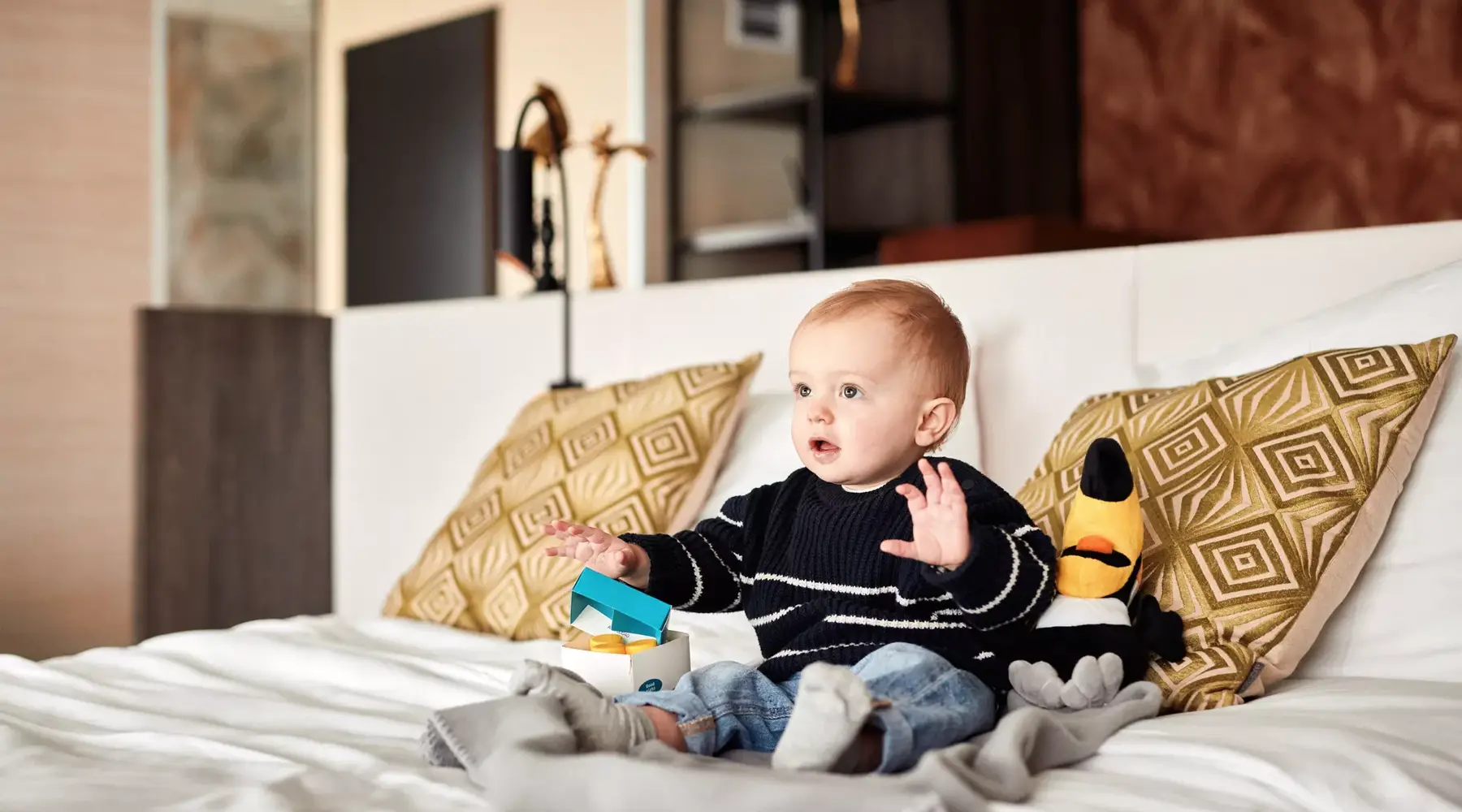 Baby on bed in the hotel room with a Timo Toucan plush toy.