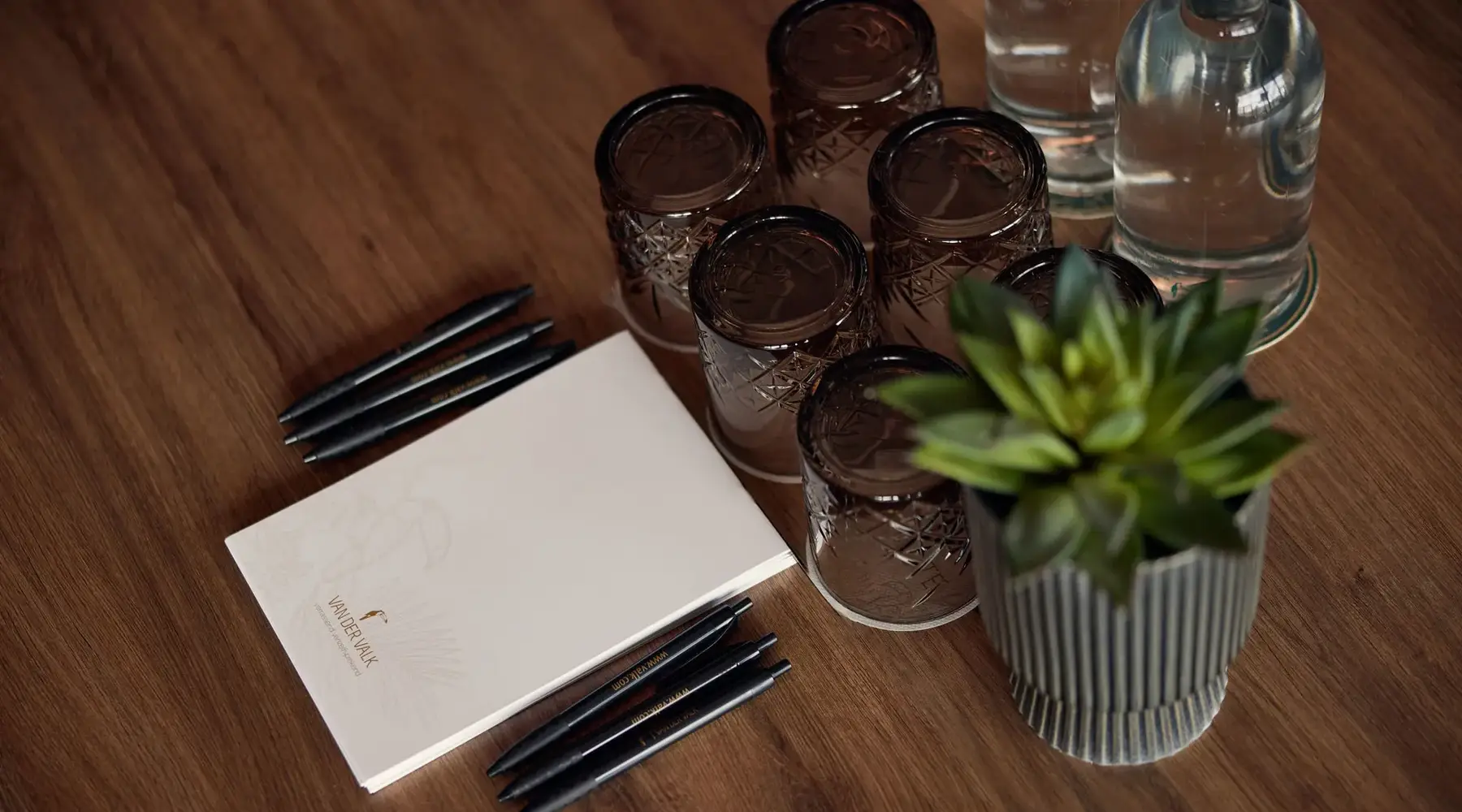 Close-up of a wooden table at the Van der Valk Hotel Hildesheim with a notebook in Van-der-Valk design and several black pens. Beside it are decorative glasses and a small plant, creating a calm, stylish working or meeting atmosphere.