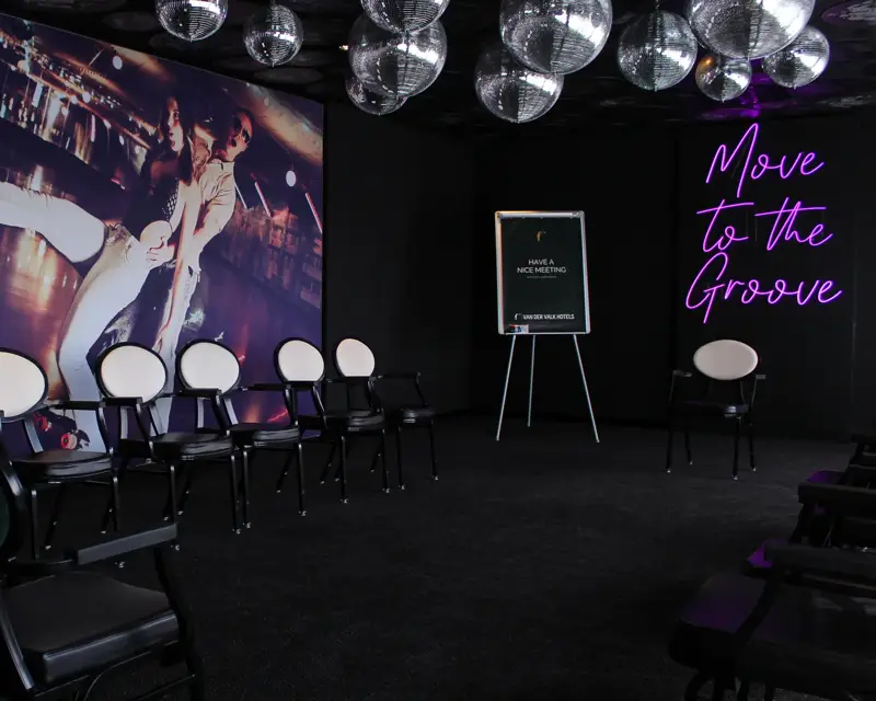 Een vergaderruimte in discostijl met discoballen aan het plafond, stoelen in opstelling en een grote wandfoto van dansers.