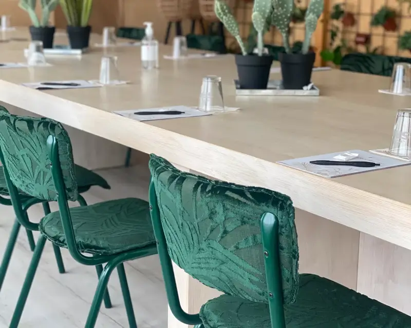 Ein Tagungsraum mit einem langen Holztisch, grünen Stühlen, Kakteen als Dekoration und einer Pflanzenwand in natürlicher Atmosphäre.