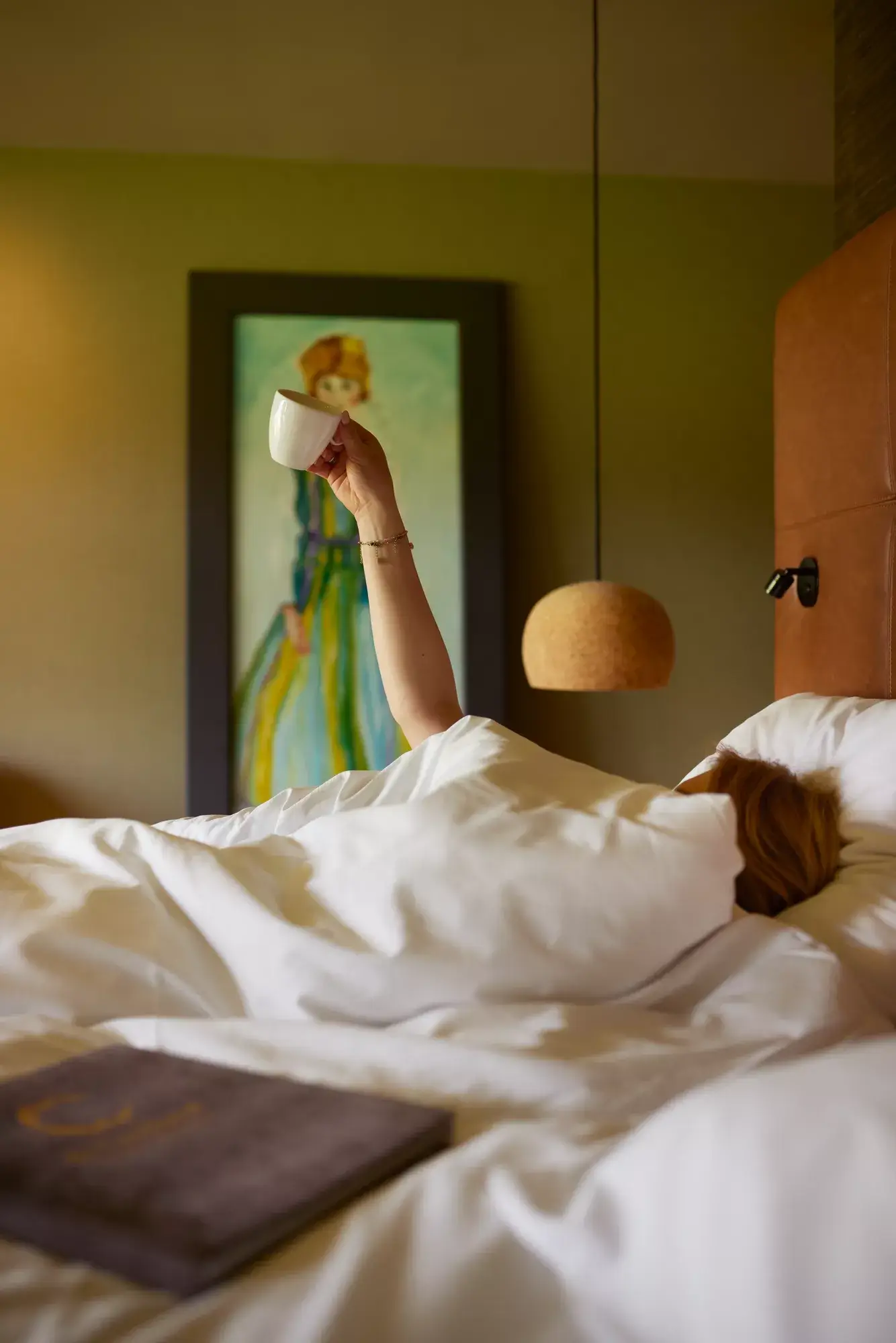 Lady in a hotel bed raising her hand with a cup of coffee