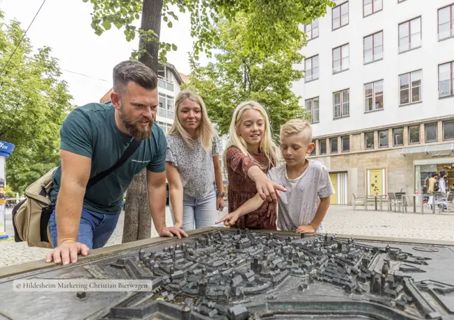 Vierköpfige Familie steht im Freien um ein detailliertes Stadtmodell aus Metall und betrachtet es neugierig; zwei Kinder zeigen auf bestimmte Stellen, während die Eltern sich interessiert darüber beugen, umgeben von Bäumen und Gebäuden in einer ruhigen Stadtumgebung.