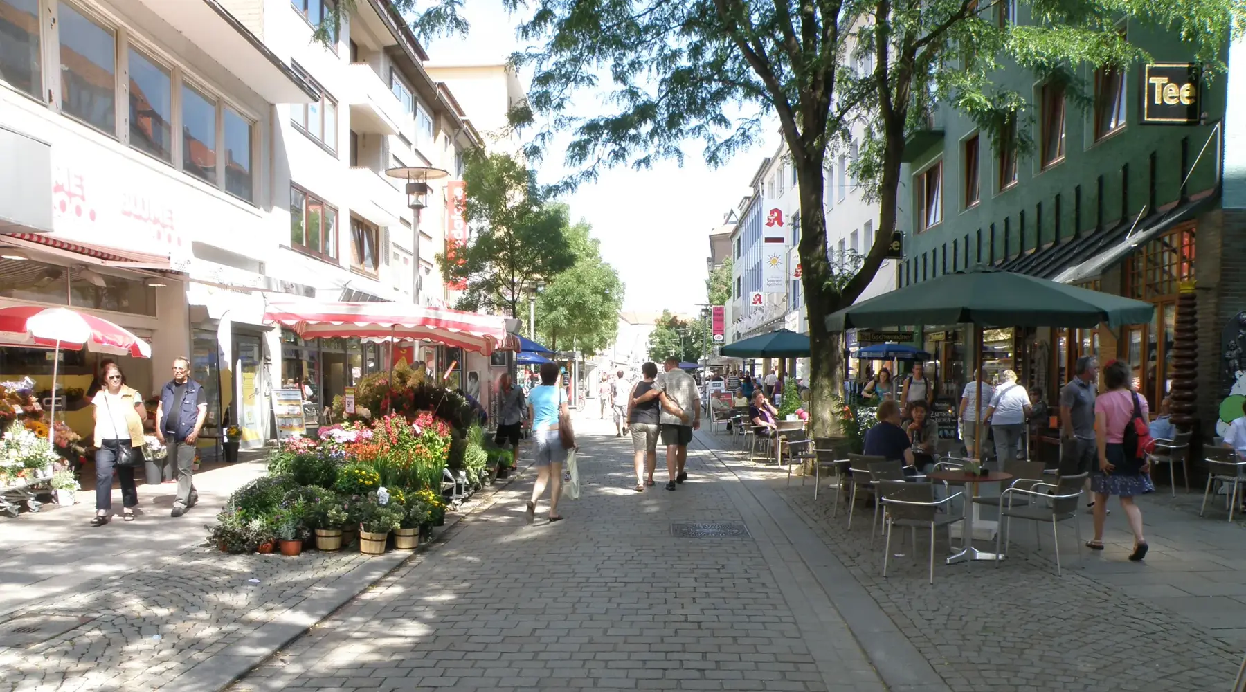 Winkelstraat in het centrum van Hildesheim met winkels, cafés en zitgelegenheden buiten. Mensen lopen door de winkelstraat of zitten aan de tafels voor de horecagelegenheden. Bloemendisplays, bomen en de geplaveide straat creëren een levendige, vriendelijke sfeer.