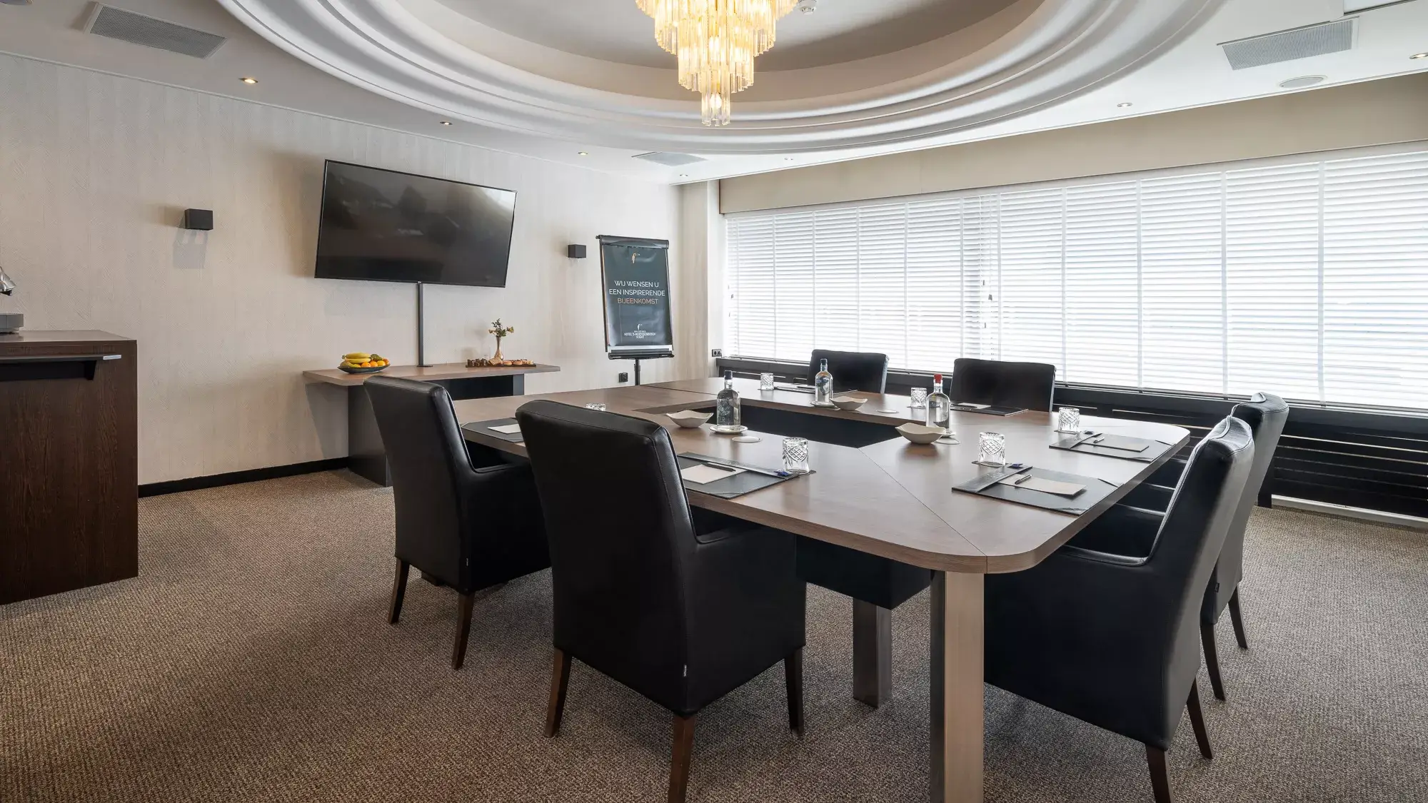 Overview of the modern Bach Room in a block configuration, equipped with comfortable meeting chairs, plenty of natural light, and an LCD screen on the wall.