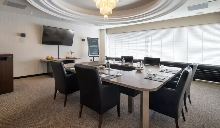 Overview of the modern Bach Room in a block configuration, equipped with comfortable meeting chairs, plenty of natural light, and an LCD screen on the wall.