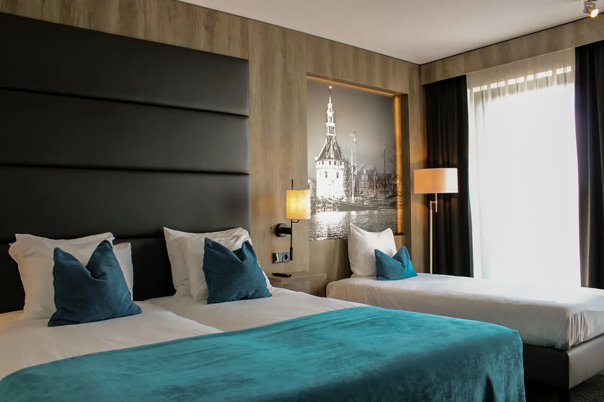 Hotel room with a double bed and a single bed, turquoise bedspread, blue decorative pillows, wooden wall finish, and a black-and-white photo of the Hoofdtoren in Hoorn.
