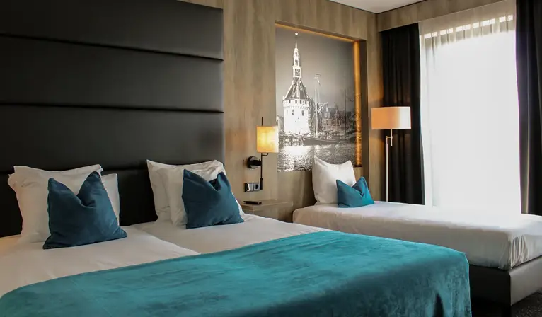 Hotel room with a double bed and a single bed, turquoise bedspread, blue decorative pillows, wooden wall finish, and a black-and-white photo of the Hoofdtoren in Hoorn.