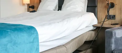 Detailansicht eines Hotelzimmers mit elektrisch verstellbarem Doppelbett, weißer Bettwäsche, türkisfarbener Tagesdecke, Wandlampen, Telefon und Steckdose neben dem Bett.